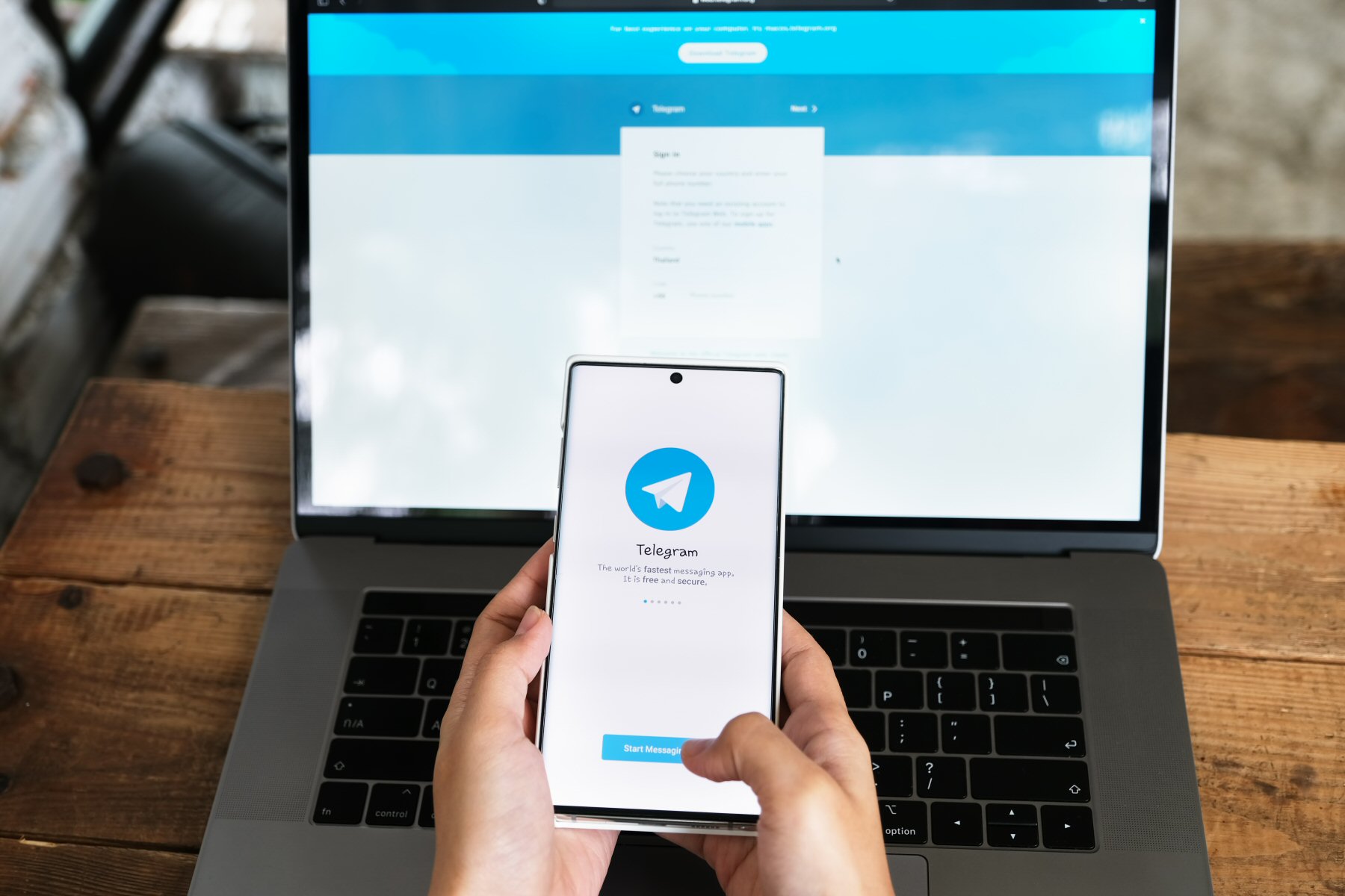 Hands holding a phone with Telegram on the screen in front of a laptop